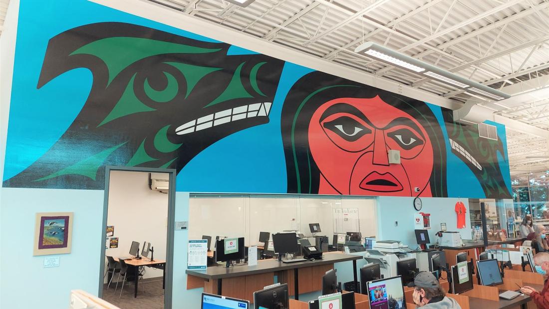 A mural inside a library above computers. An Indigenous man and two green wolves