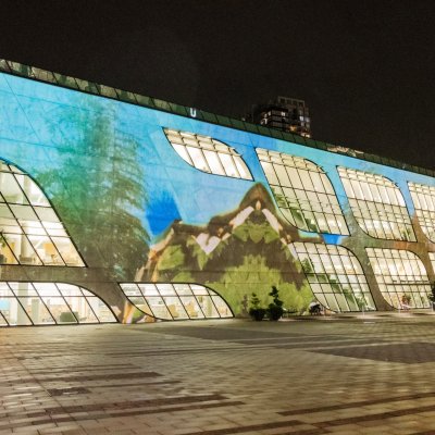 Video projection of trees, mountains, and blue sky on the facade of Surrey Central Library at nighttime.