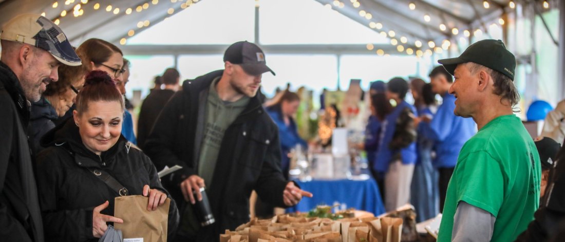 People interacting at a market stall with brown paper bags of products on display under string lights in a tented area.