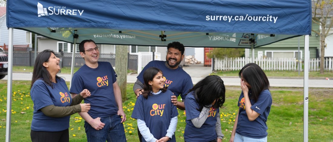 A joyful group of people wearing "Our City" t-shirts under a City of Surrey tent, sharing a laugh together at a community event.